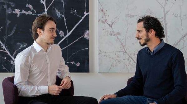 André Semadeni und Michael Naegeli sitzen während dem Interview vor zwei hängenden Bildern mit feinen Blumen.